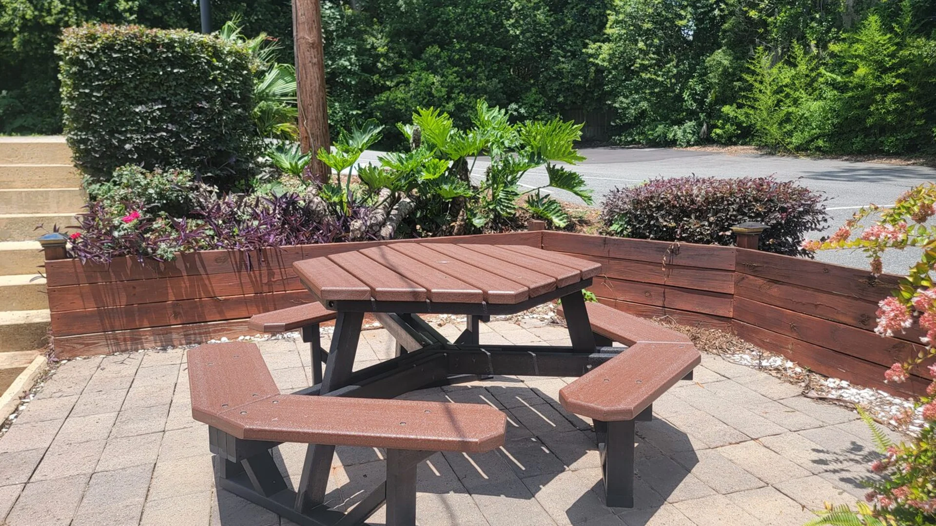 Outdoor patio area with a hexagonal picnic table surrounded by lush greenery and landscaped flower beds